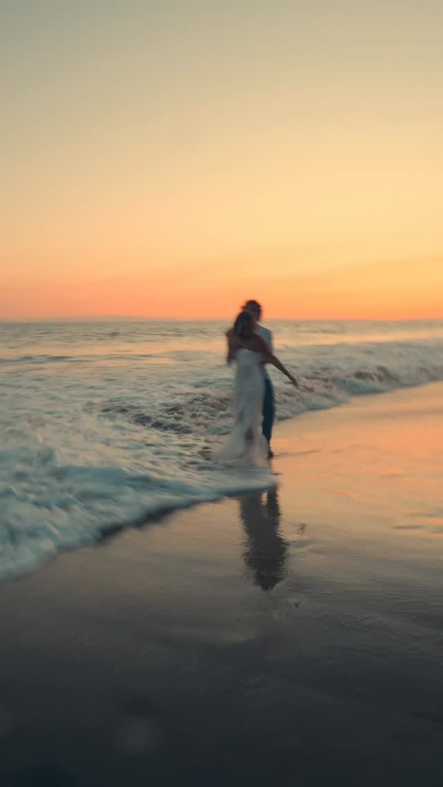 Couple standing on a beach at sunset with waves washing over their feet.