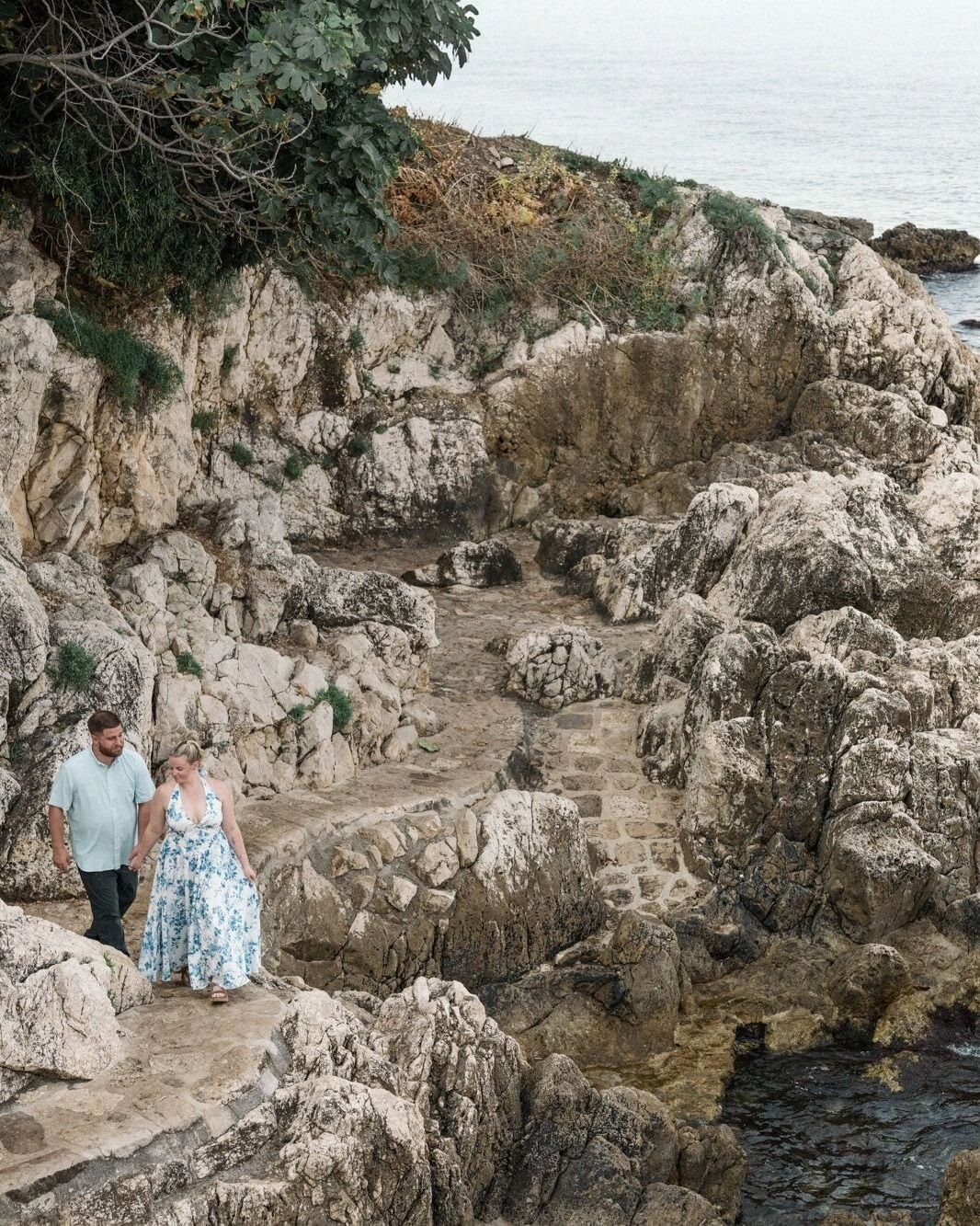 Couple walking hand-in-hand along a rugged, rocky coastal path by the ocean.