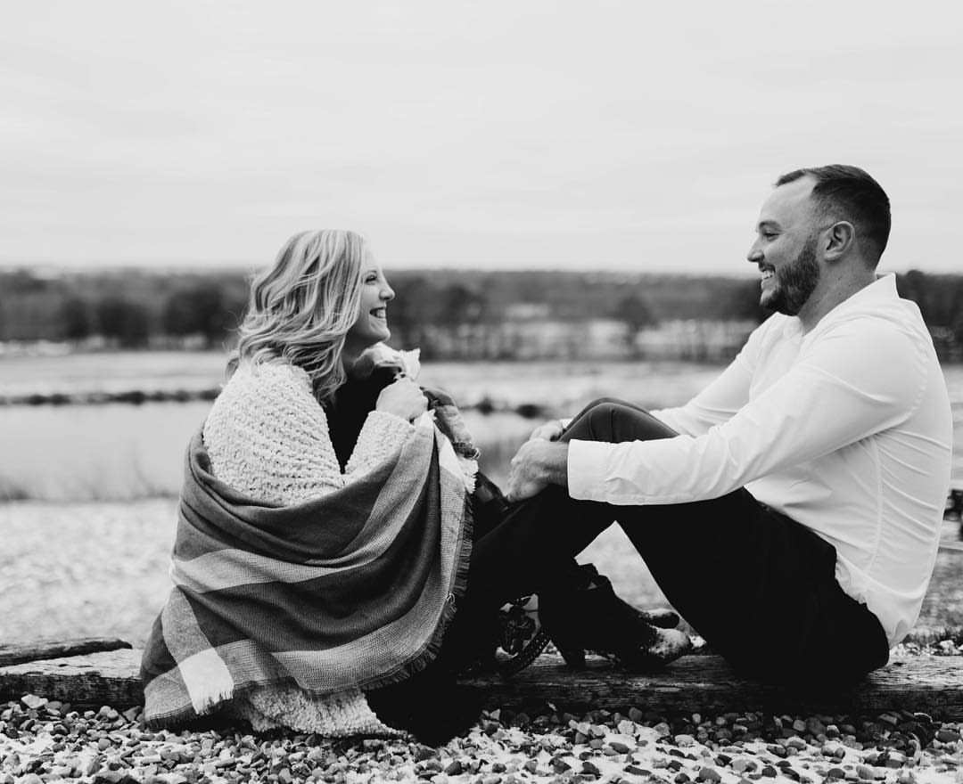 Couple sitting on a rocky beach with the woman wrapped in a cozy blanket.