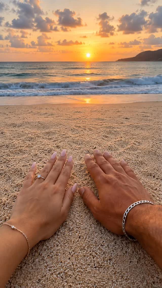 Two hands resting on beach sand with an engagement ring during a sunset.
