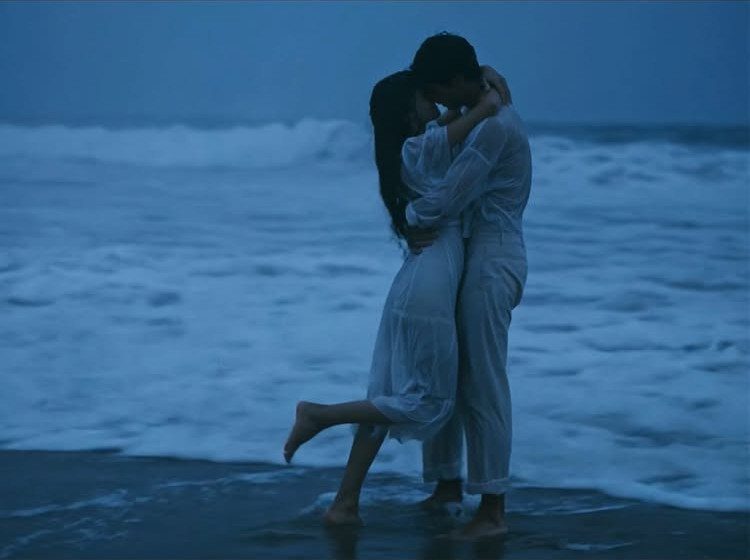 Couple in white clothing kissing while standing in shallow ocean waves at twilight.