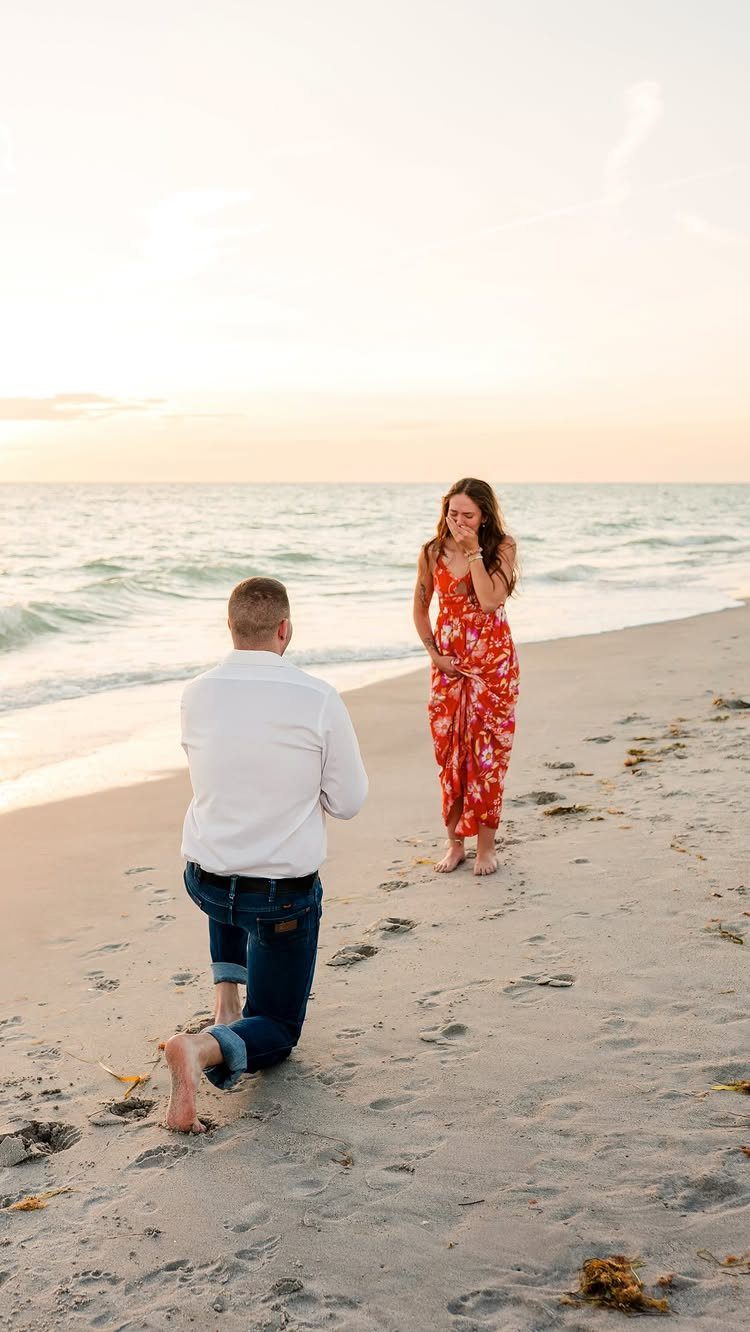 Man kneeling on a sandy beach proposing to a woman in a red dress.