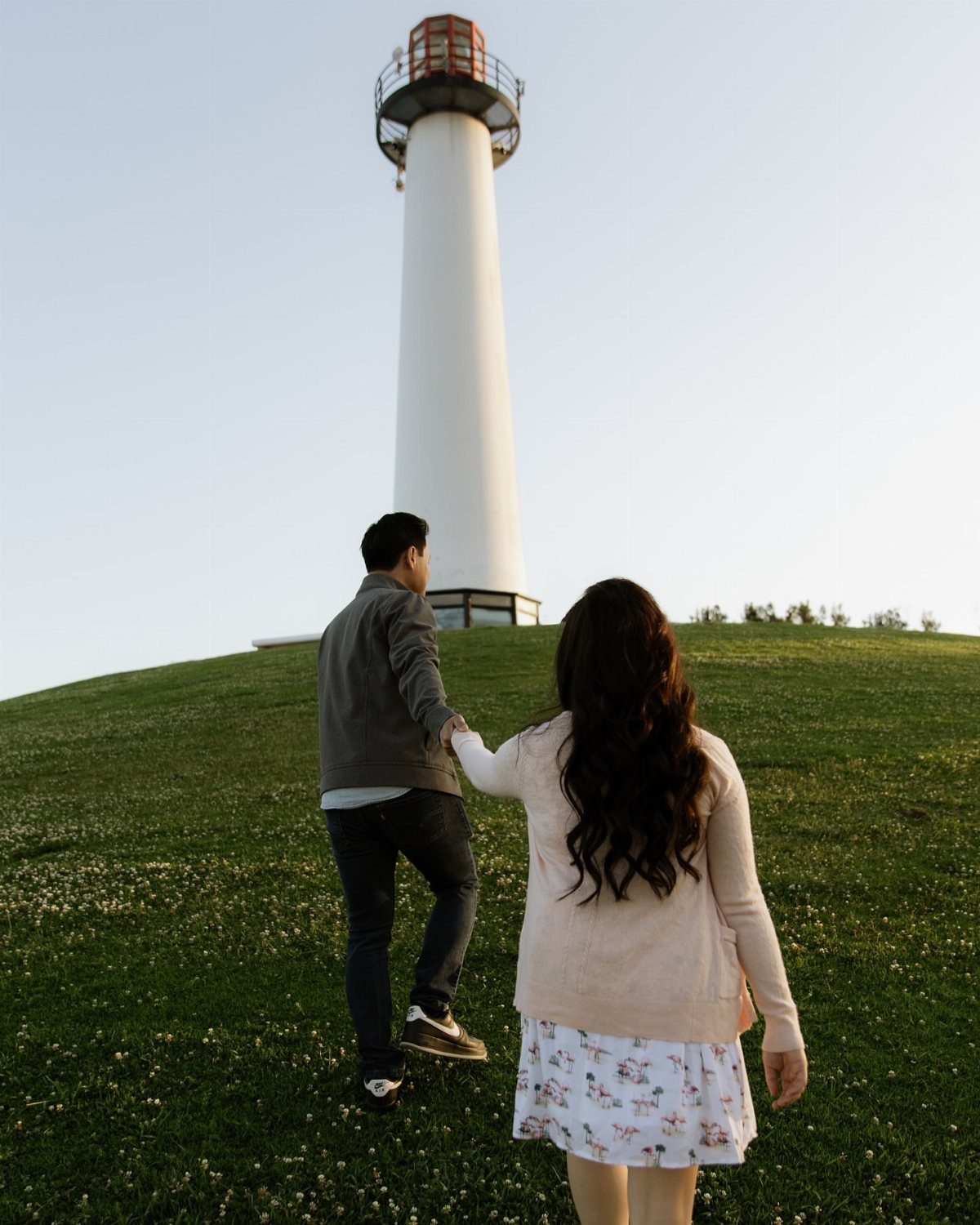 Couple holding hands walking up a grassy hill toward a white lighthouse.