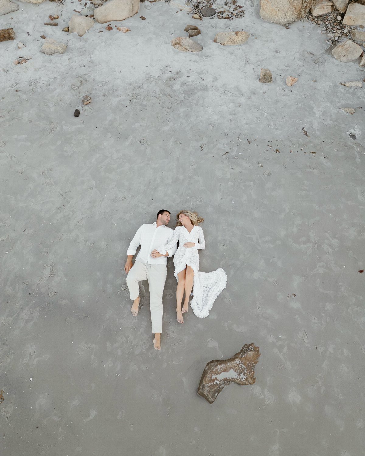 Couple in white clothing lying side-by-side on a light grey sandy beach.