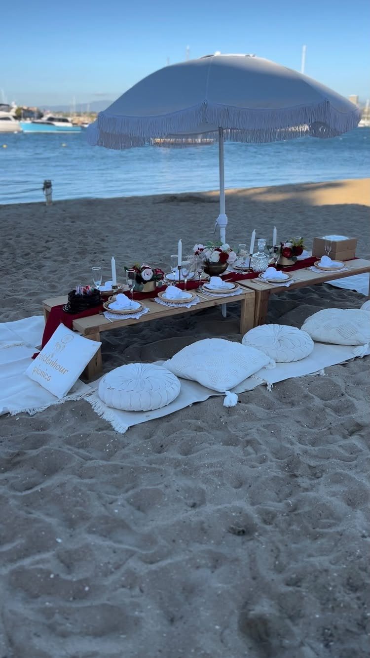 Low wooden picnic table with white cushions and umbrella on a sandy beach.