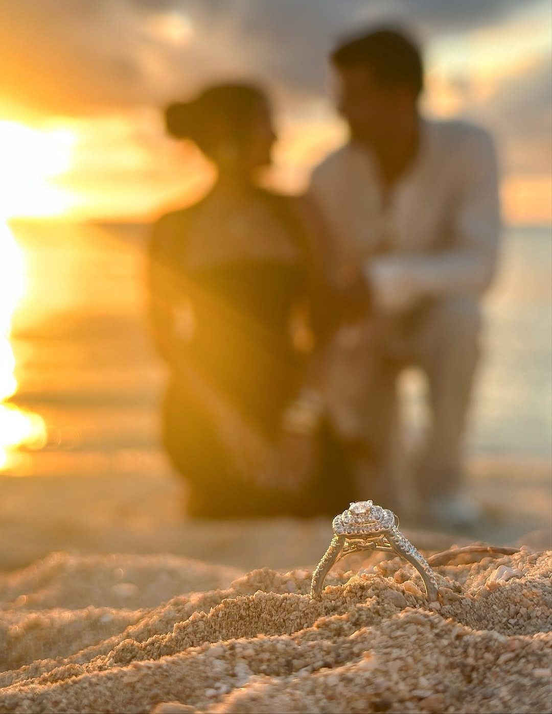 Diamond engagement ring in the sand with a blurred couple at sunset beach.
