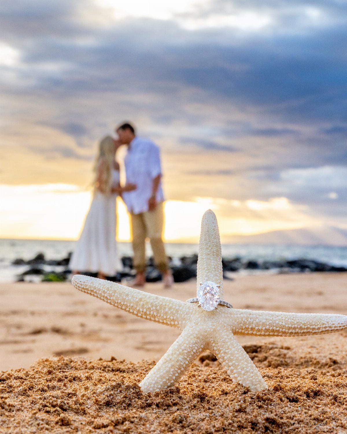 Diamond engagement ring on a white starfish with a kissing couple blurred behind.