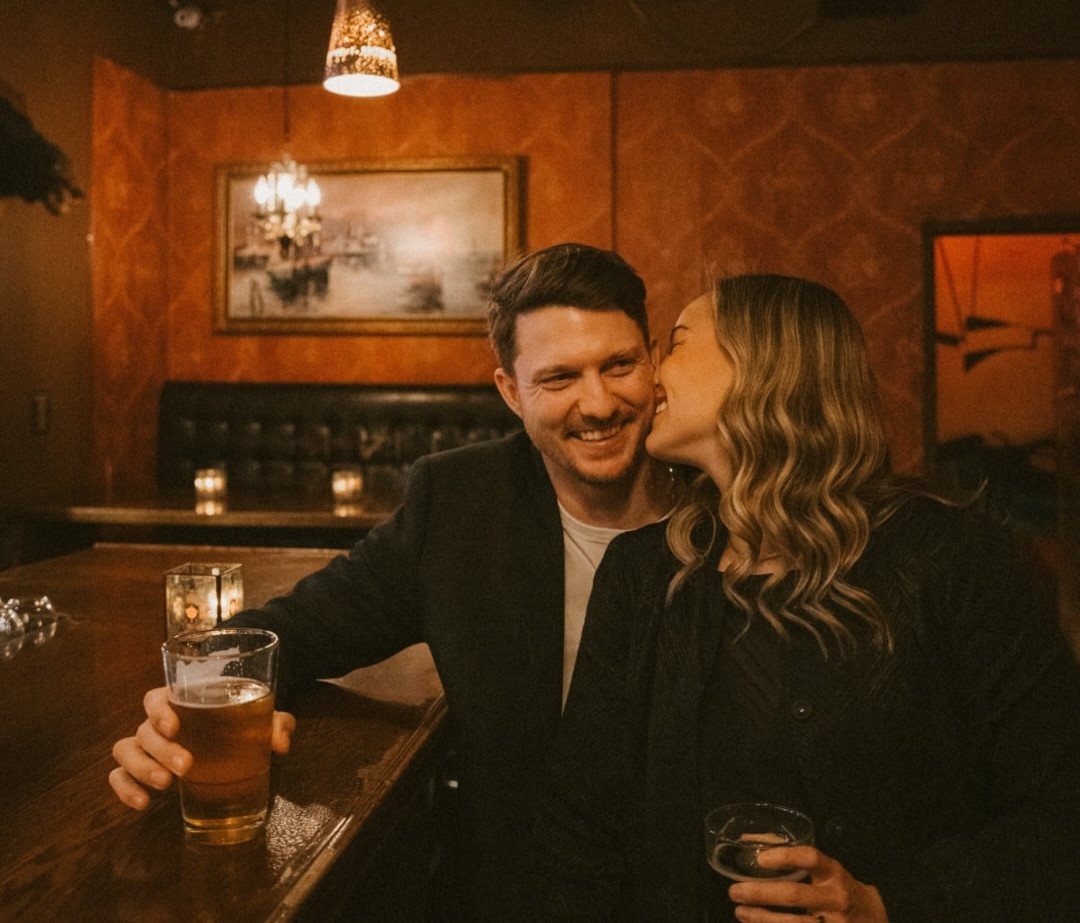 Smiling couple in casual dark clothing posing at a dimly lit wooden bar.