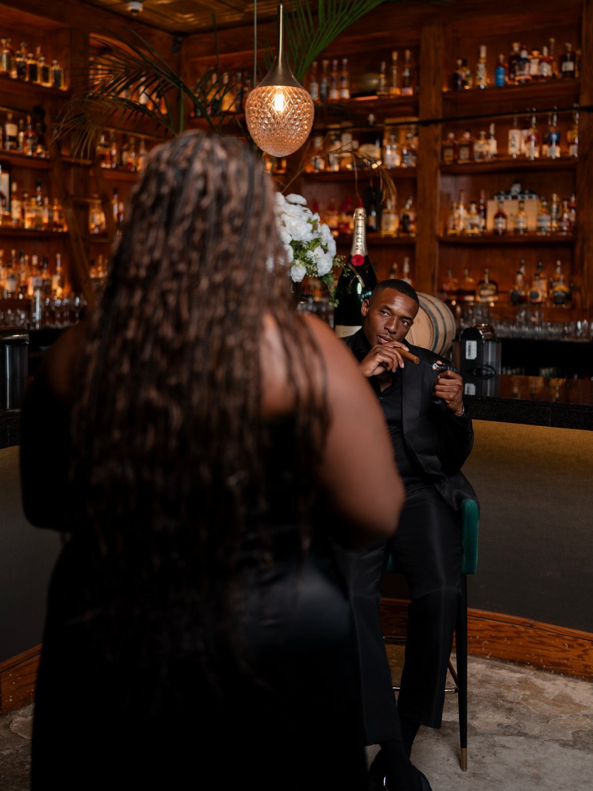 Couple in formal black attire posing in a dimly lit bar with liquor bottles.