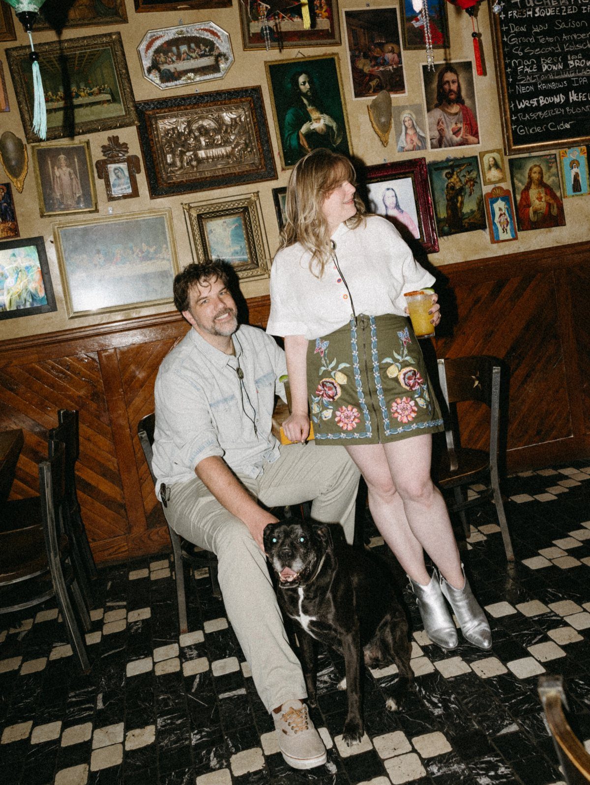 Couple and their black dog posing in a dive bar with religious art.