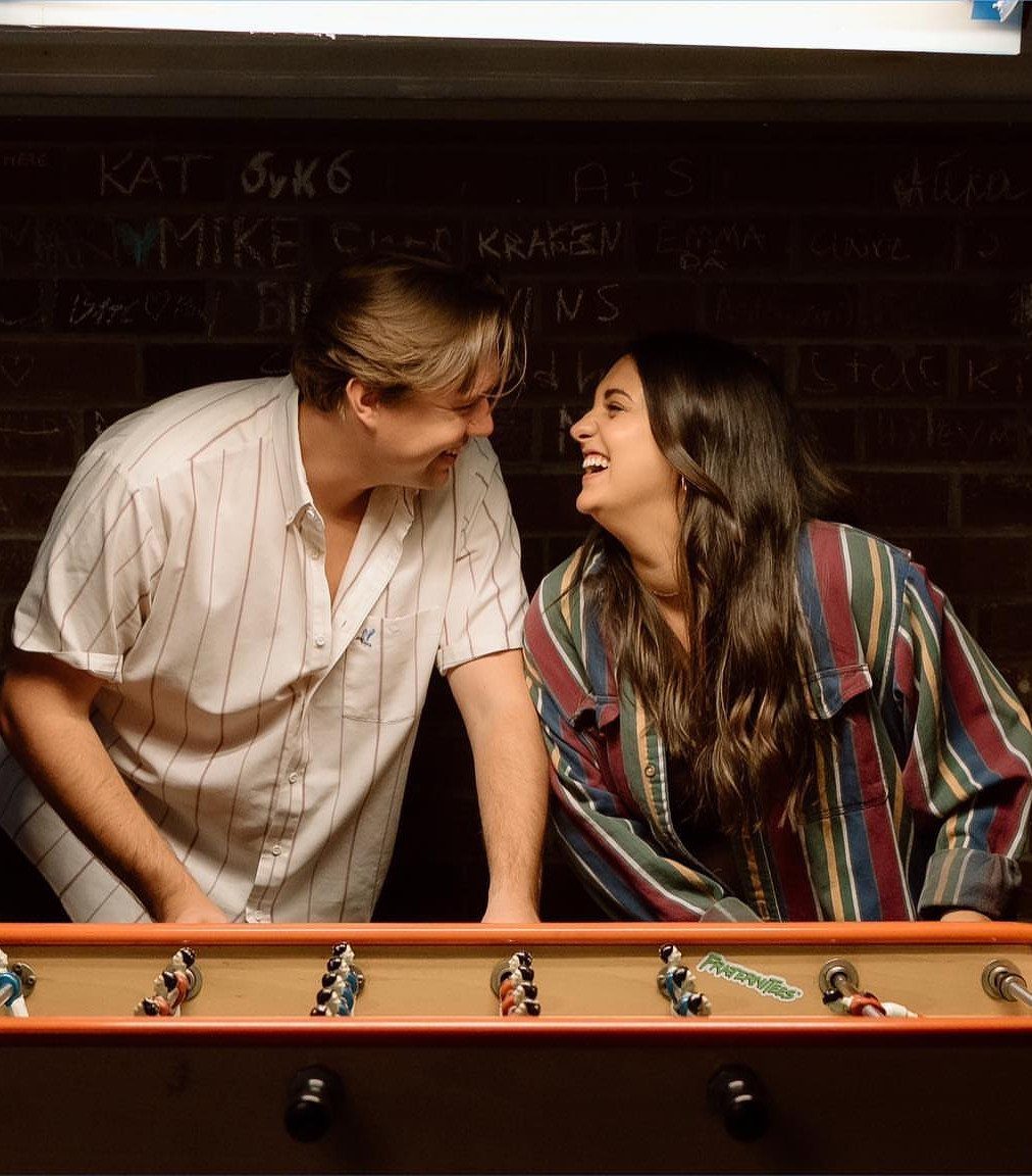 Couple smiling at each other across a foosball table in a dive bar.