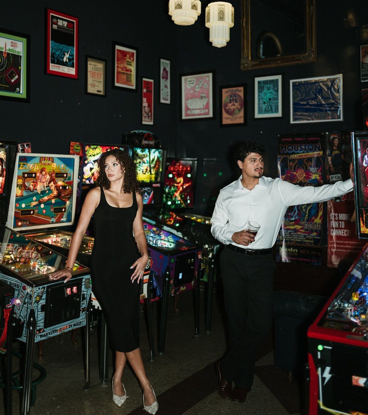 Couple posing beside colorful vintage pinball machines in a dimly lit arcade bar.