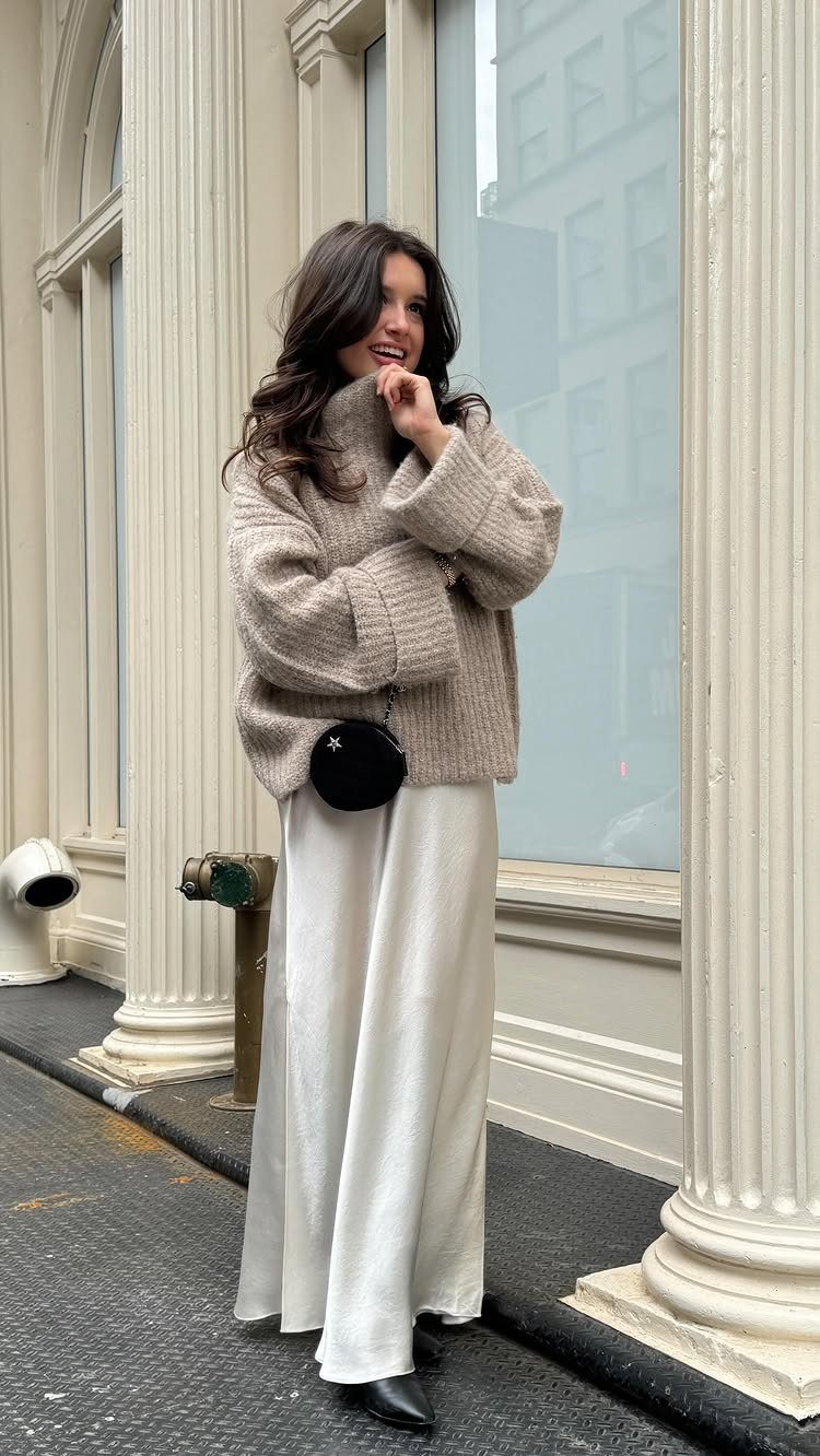 Woman in a chunky beige sweater and long white satin skirt standing outdoors.