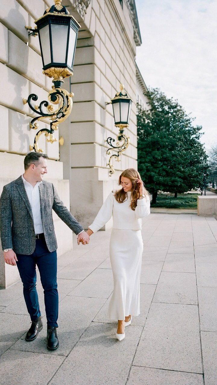 Couple holding hands walking by a building, woman wearing a white dress.