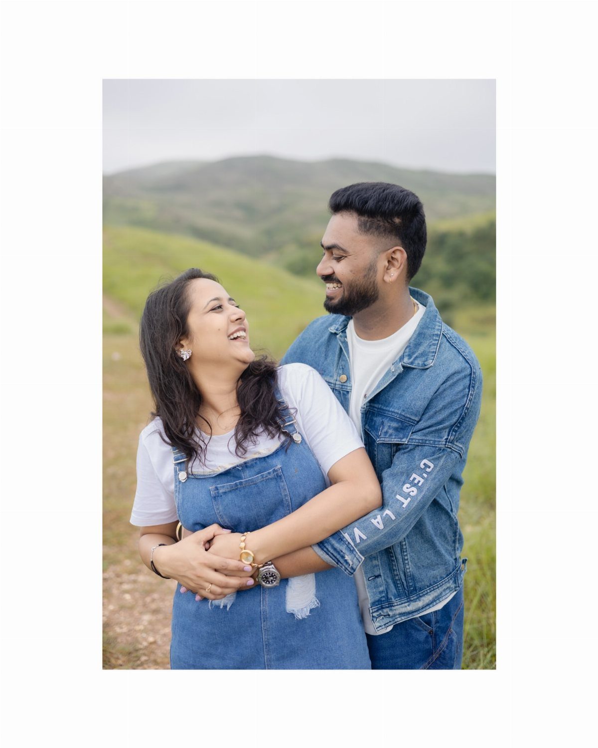 Smiling couple embracing in a grassy field with rolling green hills behind them.