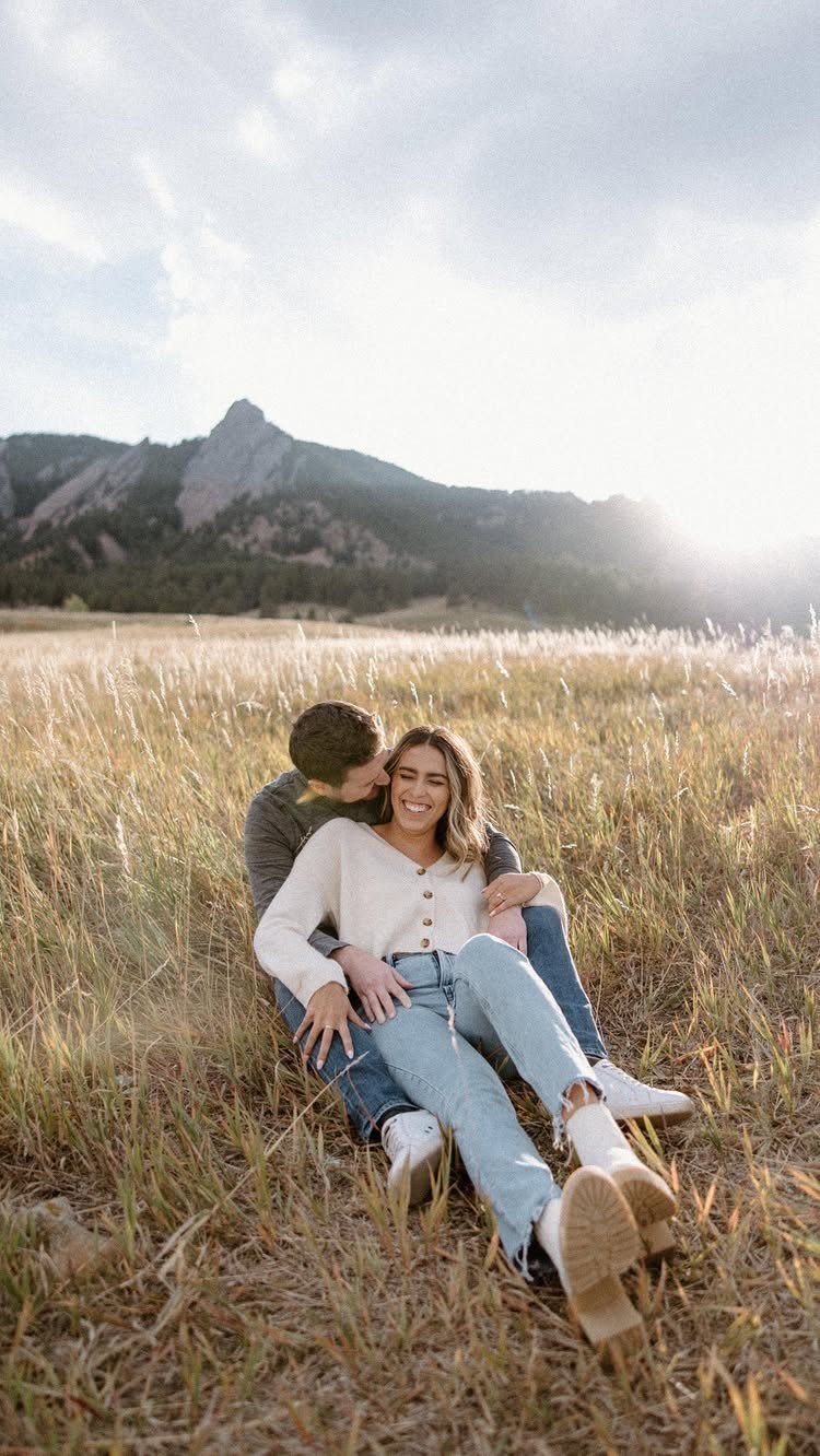 Couple sitting in golden grass wearing casual sweaters and jeans with mountains behind them