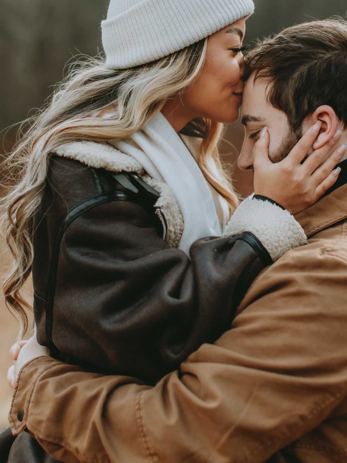 Couple in fall attire embracing, woman in white beanie kissing man's forehead.