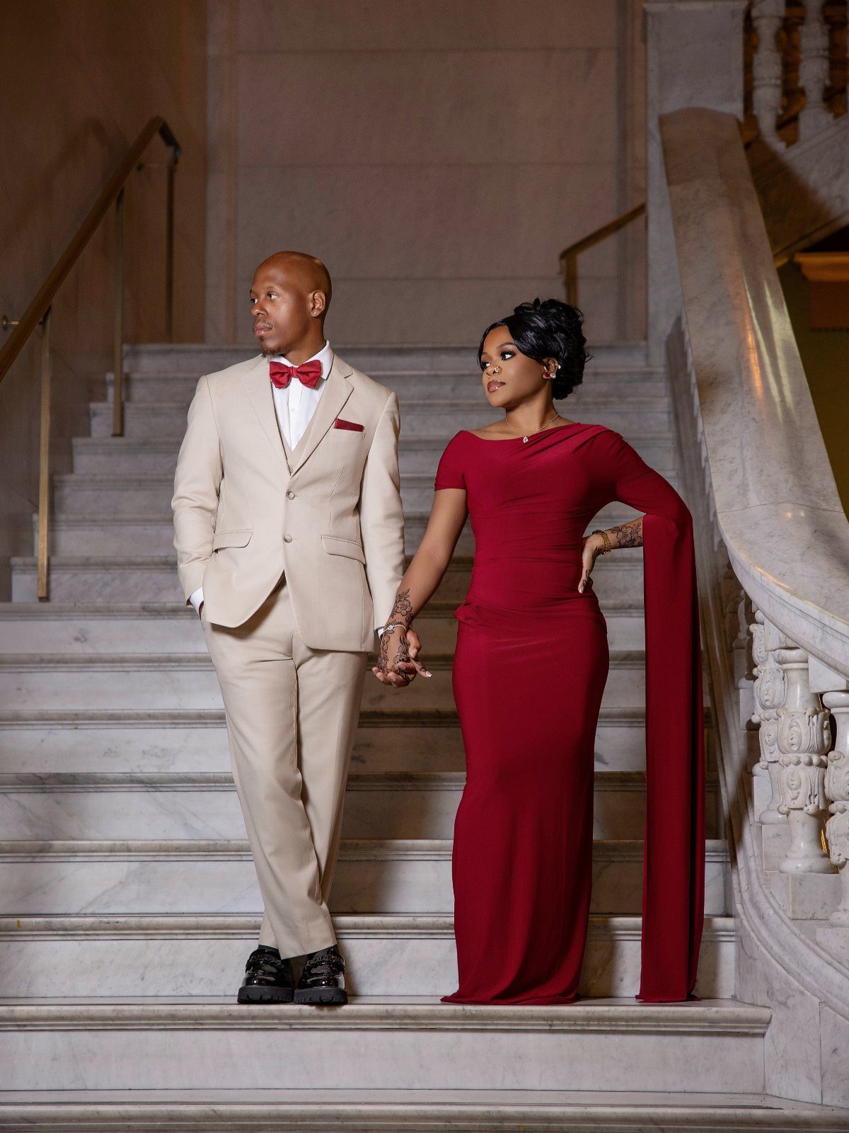 Couple in formal attire, a beige suit and red gown, on marble stairs.