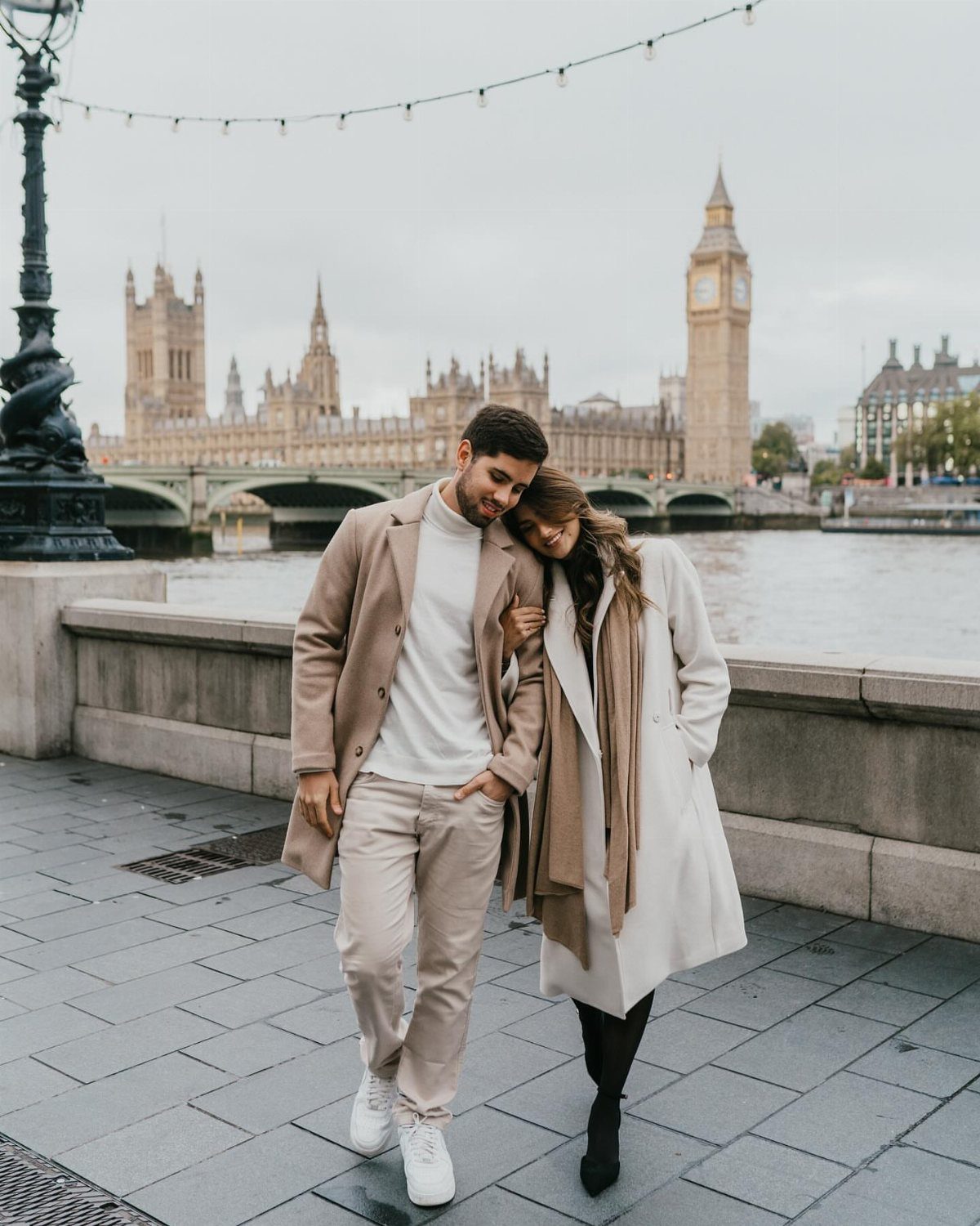 Couple in long coats posing on a London sidewalk with Big Ben behind them.