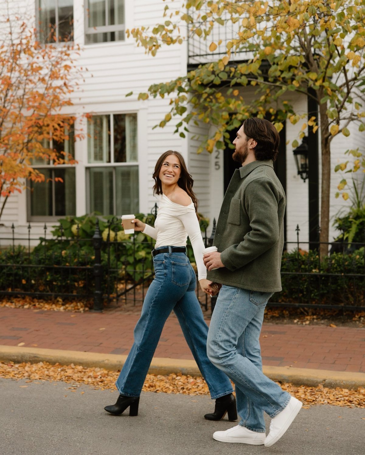 Couple walking hand-in-hand in autumn outfits on a sidewalk with fallen leaves.