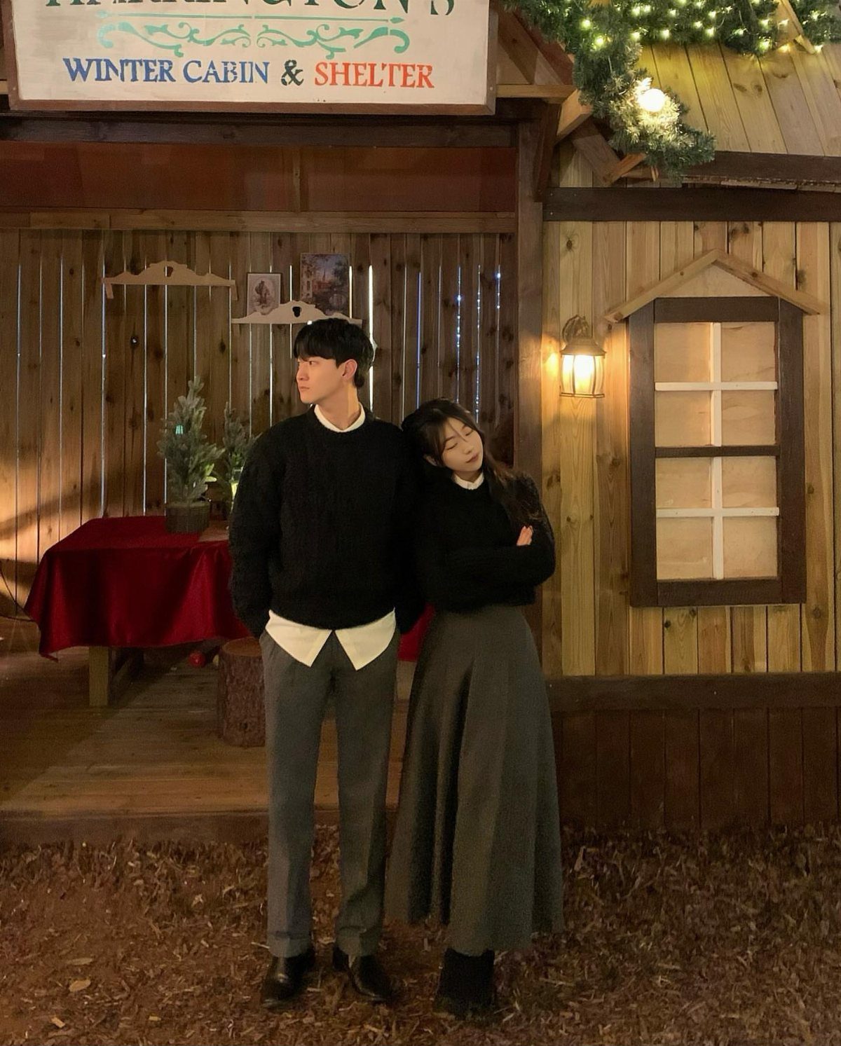 Couple in black sweaters and grey bottoms posing before a wooden winter cabin.