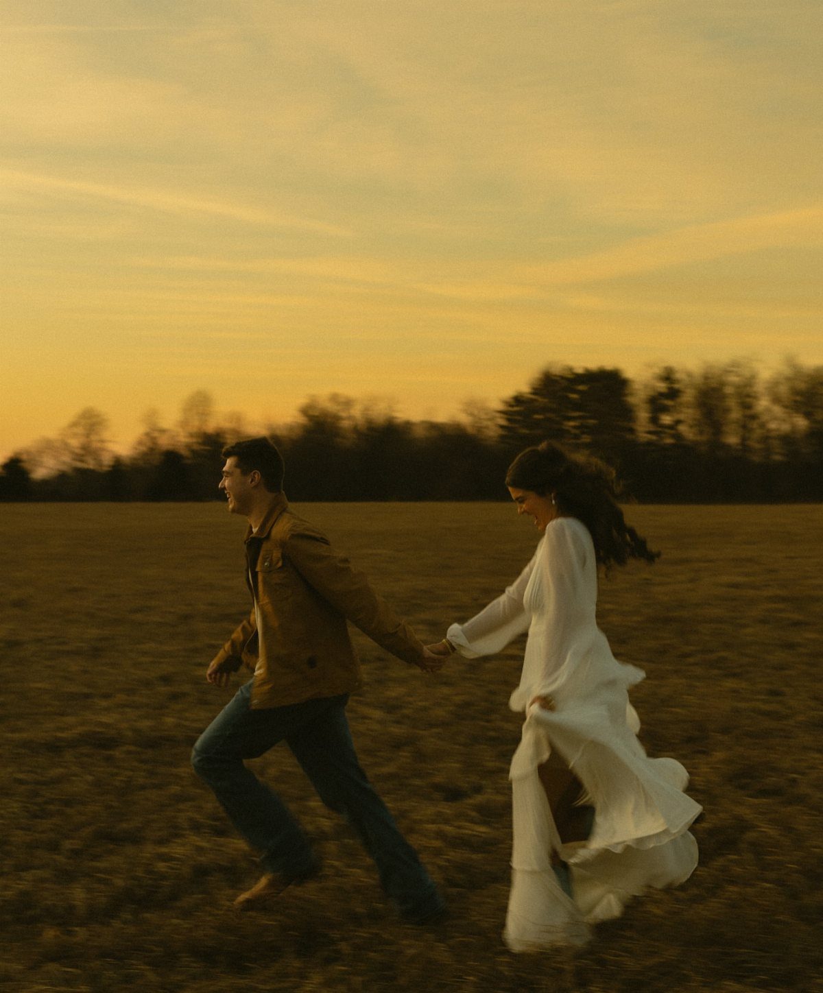 Couple running through a golden field at sunset wearing fall engagement outfits.
