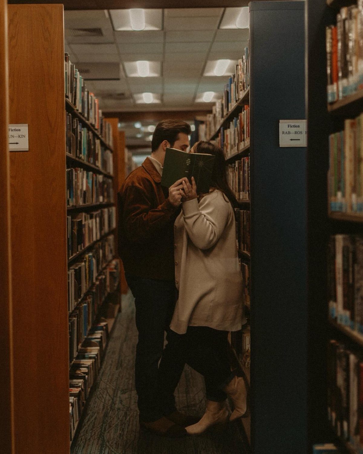 Couple holding an open green book between their faces in a library aisle