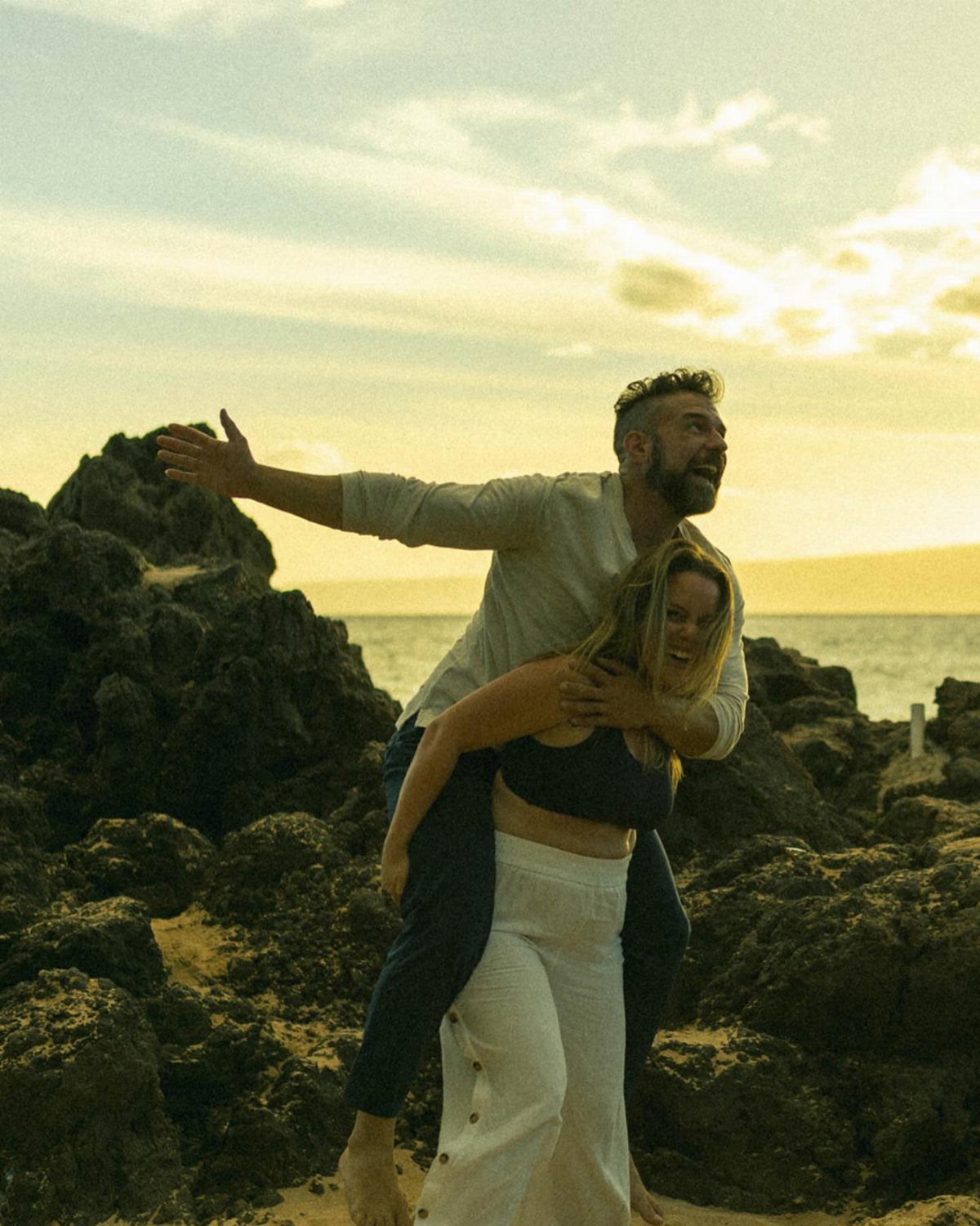 Man giving a woman a piggyback ride on a rocky beach at sunset.