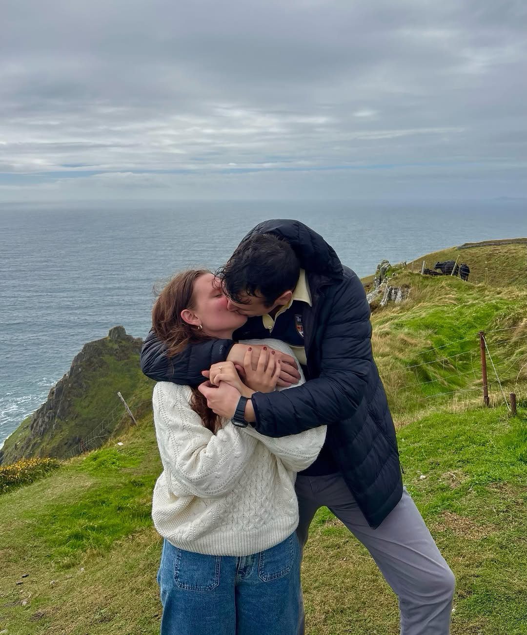 Couple kissing and embracing on a green cliff overlooking the ocean under cloudy skies.