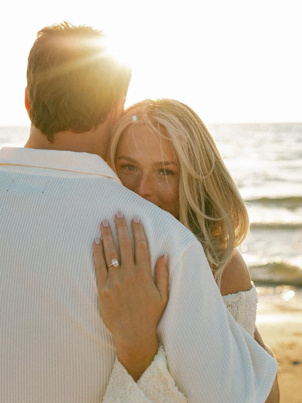 Woman embracing man from behind on a beach, showing her engagement ring.