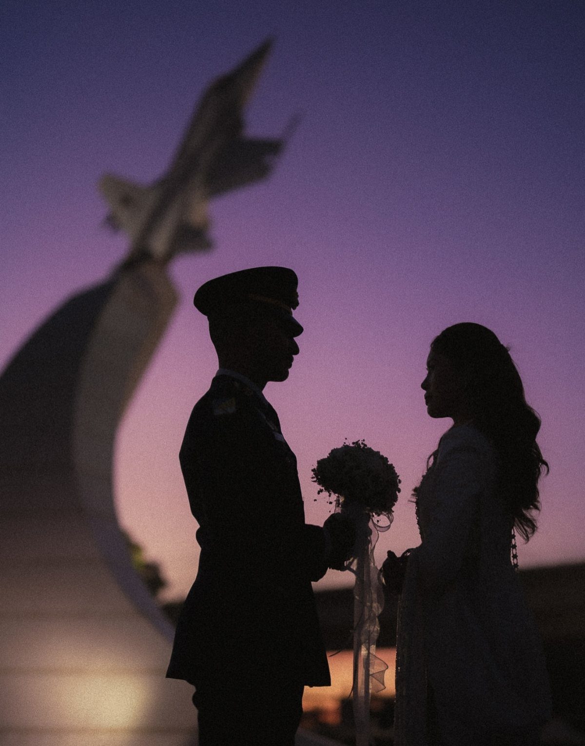 Silhouetted couple facing each other during a purple and orange sunset by a monument.