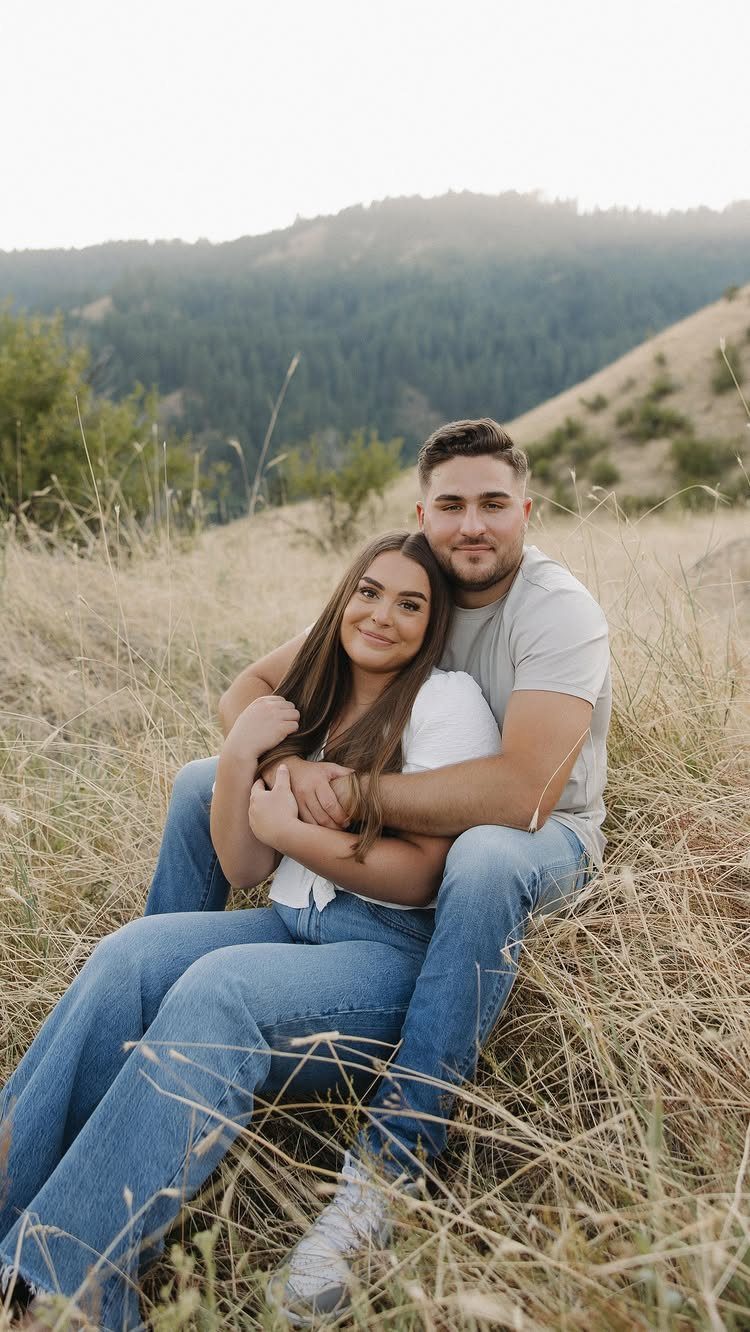 Couple sitting together in a grassy field with hills in the background.