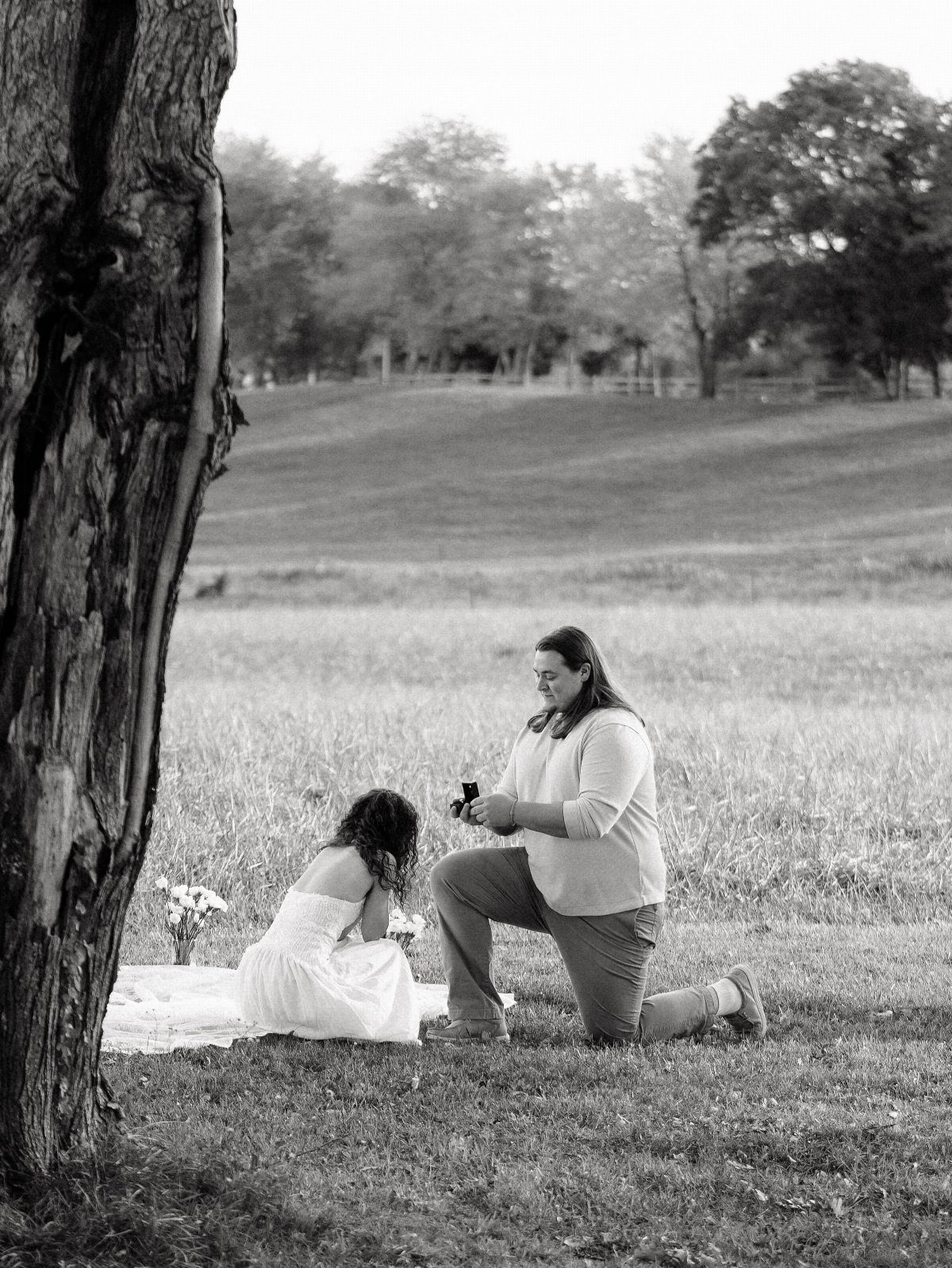 Man kneeling to propose to a woman sitting on a blanket in a field.