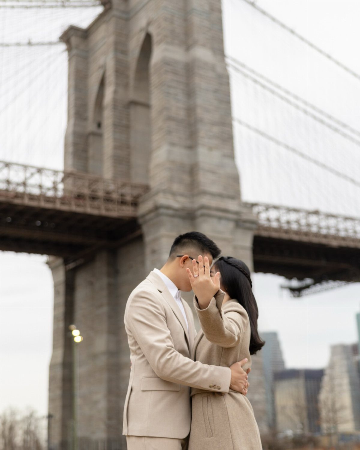Couple in beige outfits embracing and kissing in front of the Brooklyn Bridge