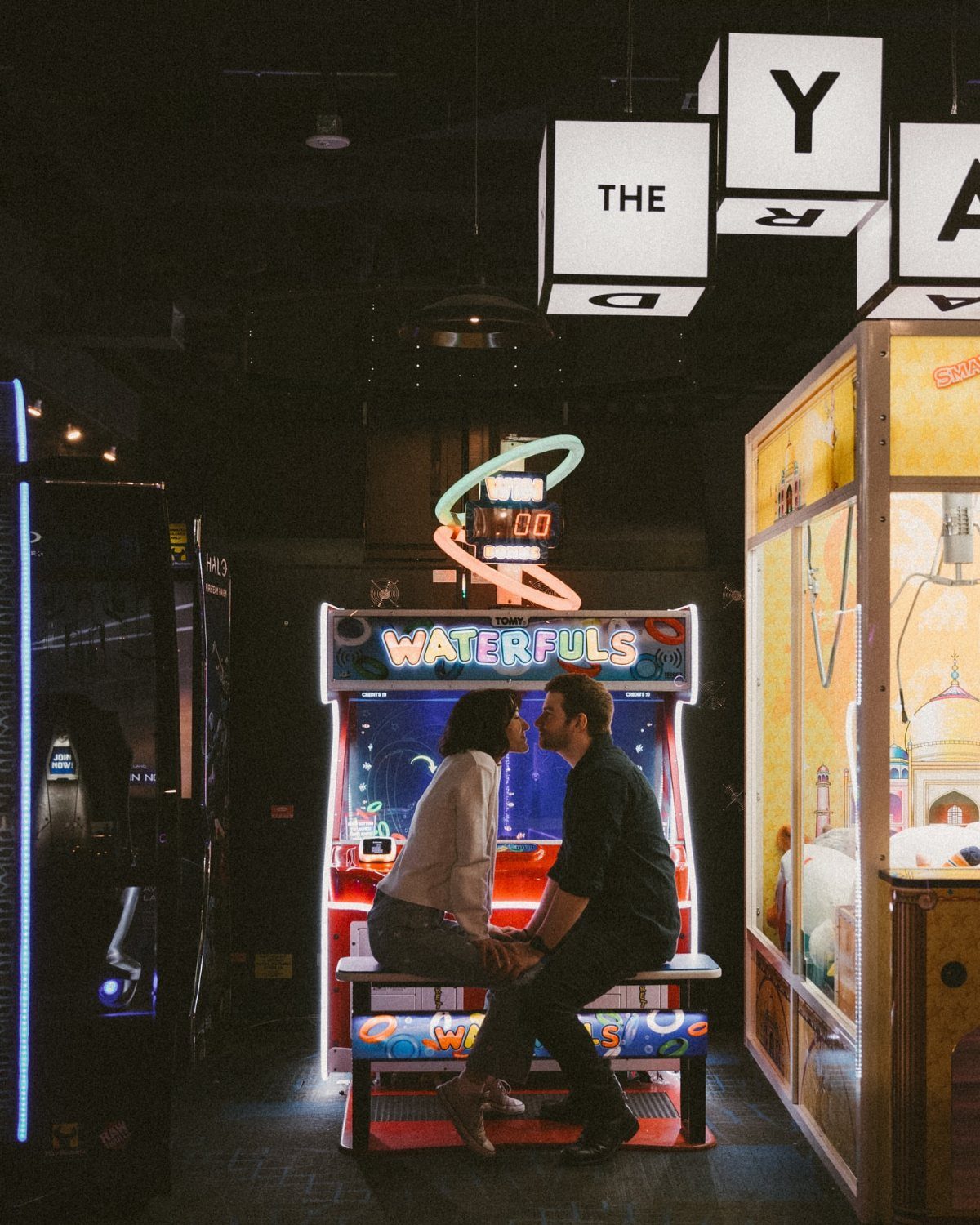 Couple sitting facing each other in front of a glowing arcade game machine.