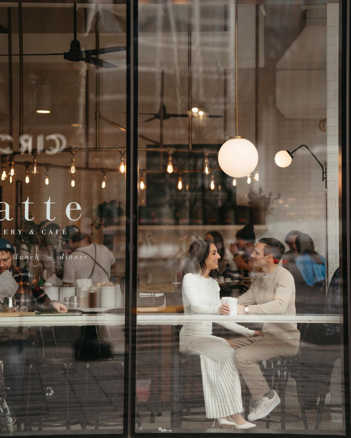 Couple sitting at a cafe counter seen through a window with warm lighting.