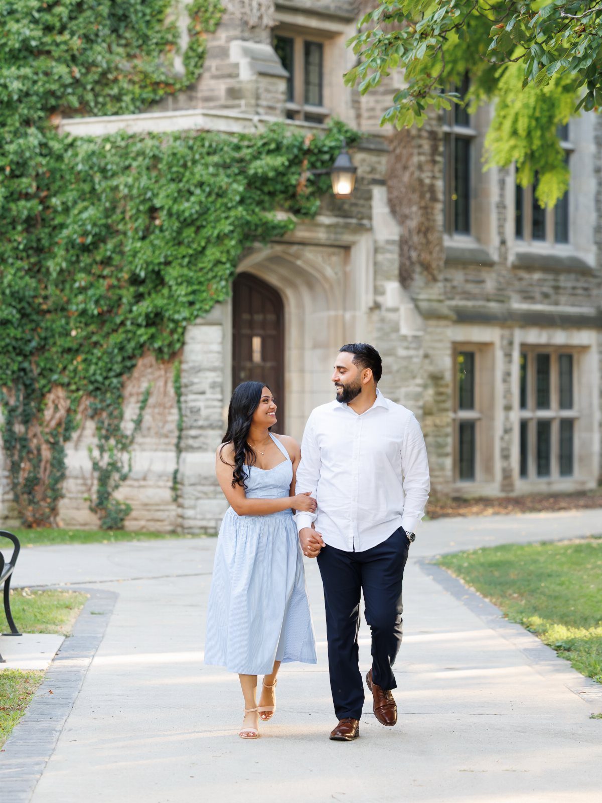 Smiling couple walking hand-in-hand on a path in front of an ivy-covered stone building