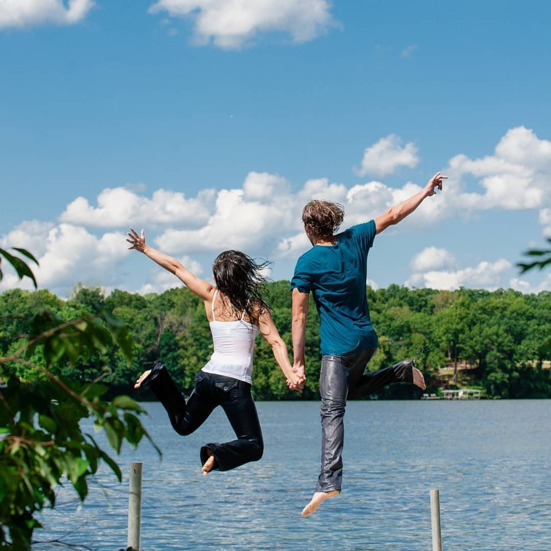 Couple jumping in the air holding hands over a lake with green trees.