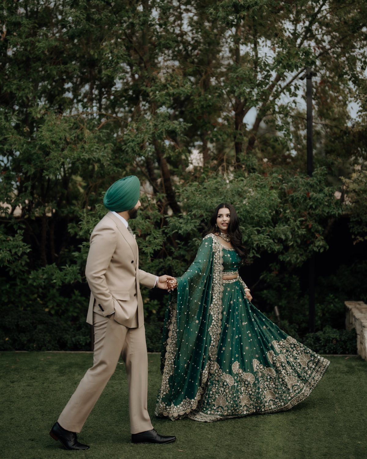 Couple holding hands, woman in a vibrant teal embroidered lehenga and man in tan.