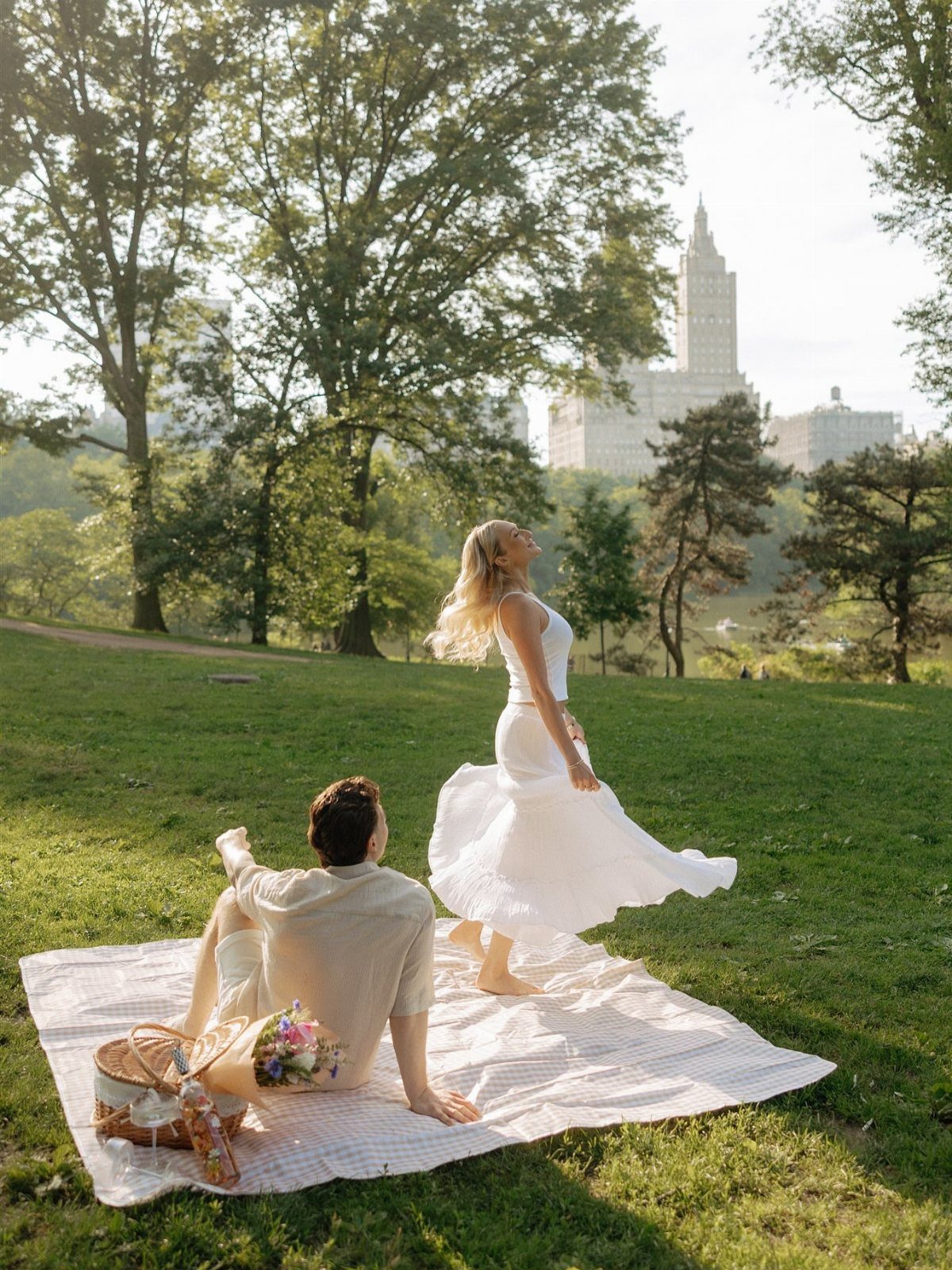 Couple on a picnic blanket in a park with a woman twirling in white.