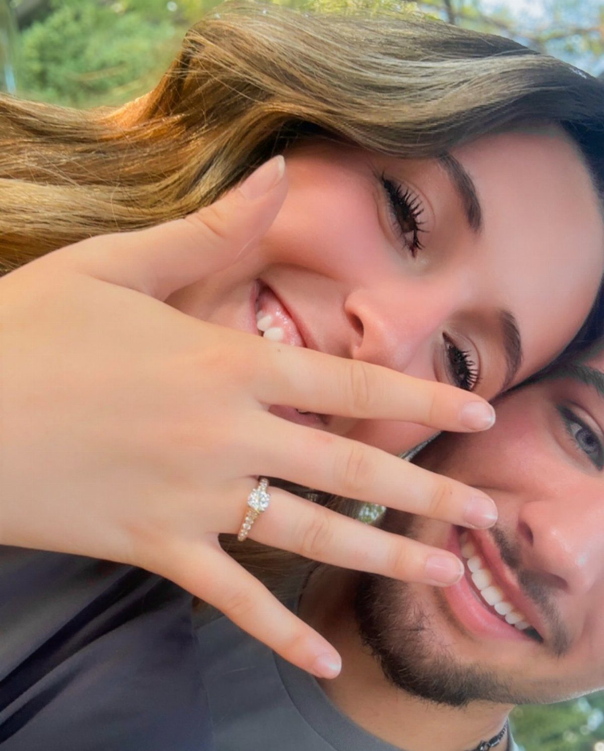 Smiling couple posing closely with a diamond engagement ring prominently displayed on a finger.