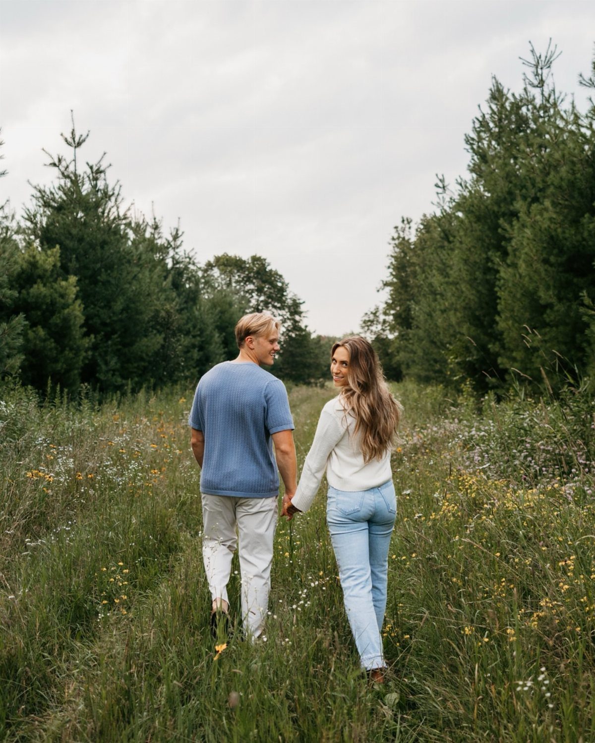 Couple holding hands while walking through a scenic grassy field surrounded by trees.