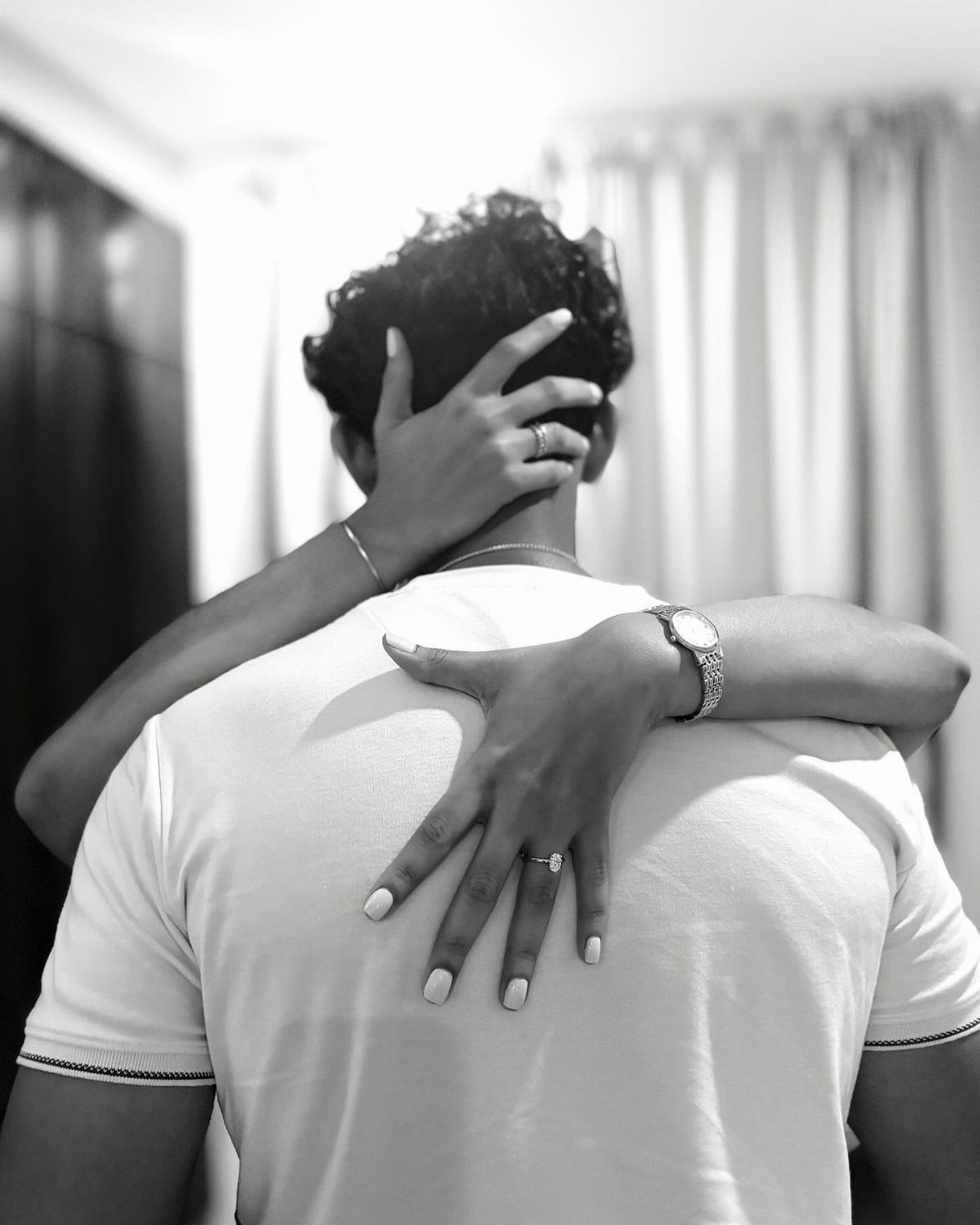 Woman hugging a man from behind, showing engagement rings on her fingers.