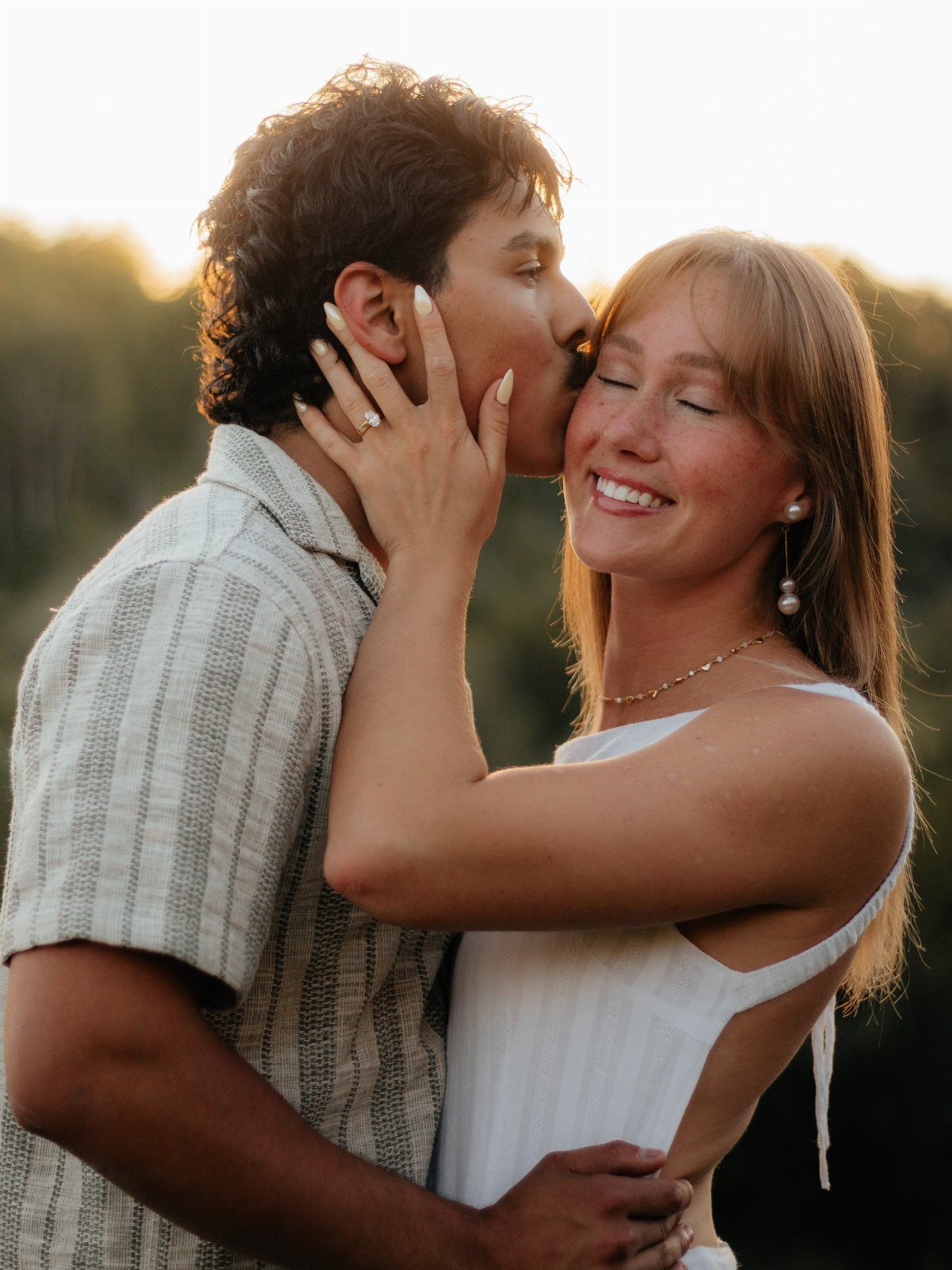 Man kissing a smiling woman on the cheek during an outdoor engagement shoot.