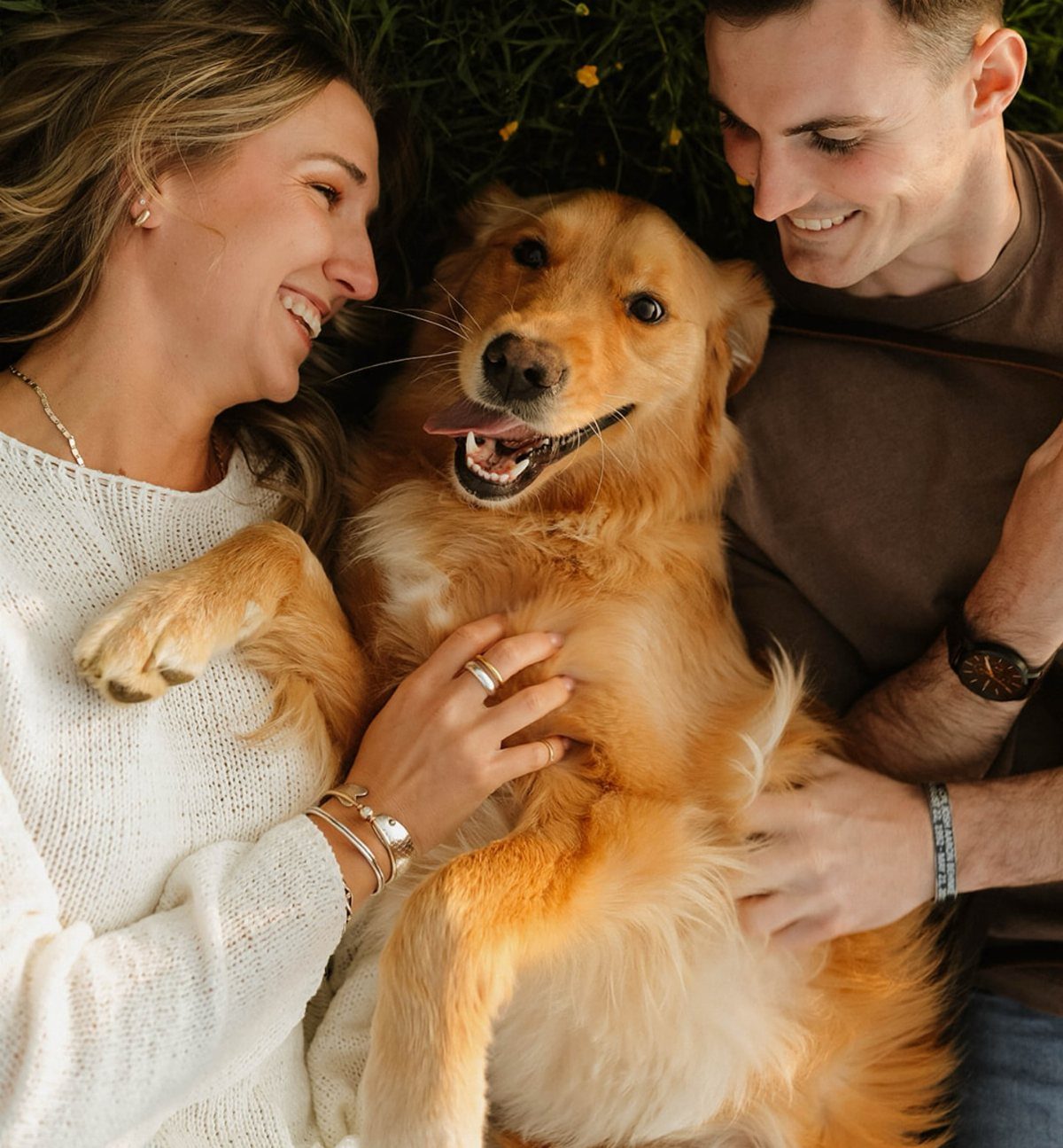Couple lying on grass cuddling a happy golden retriever during an engagement shoot.