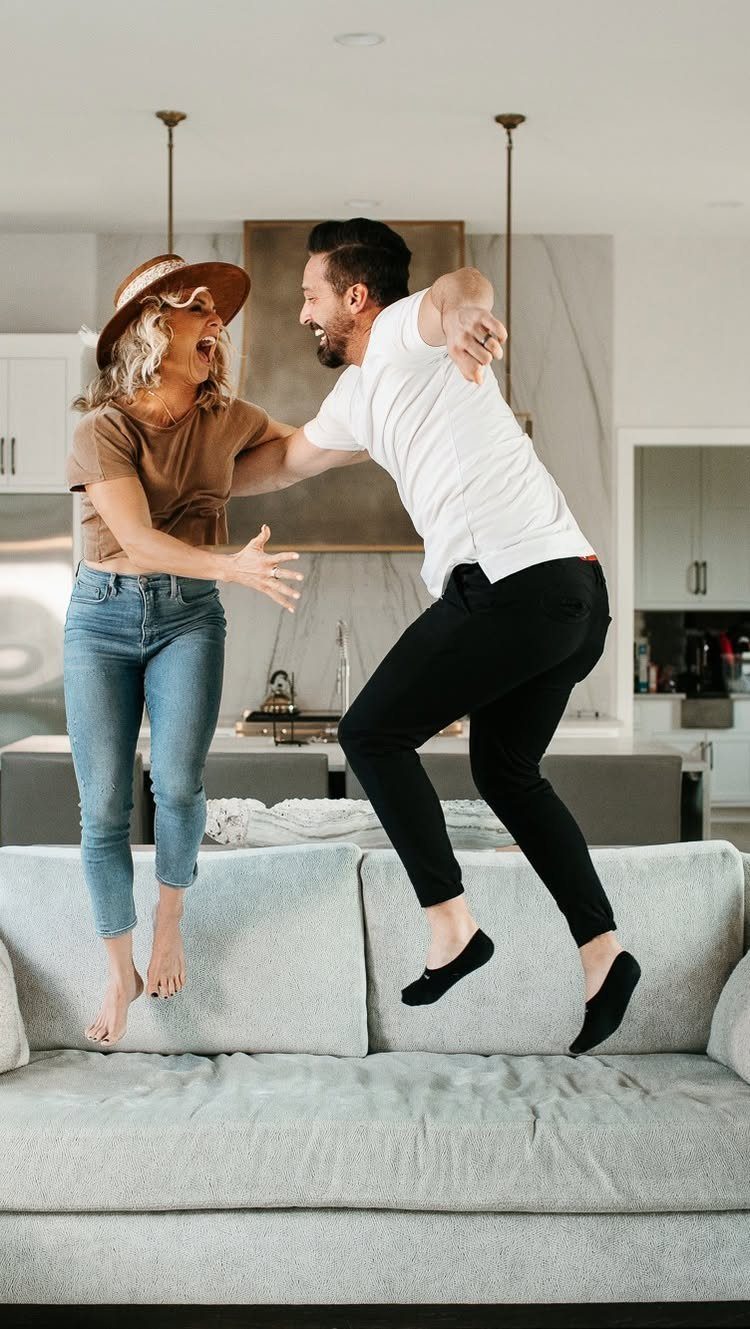 Man and woman jumping on a grey sofa in a modern white kitchen.