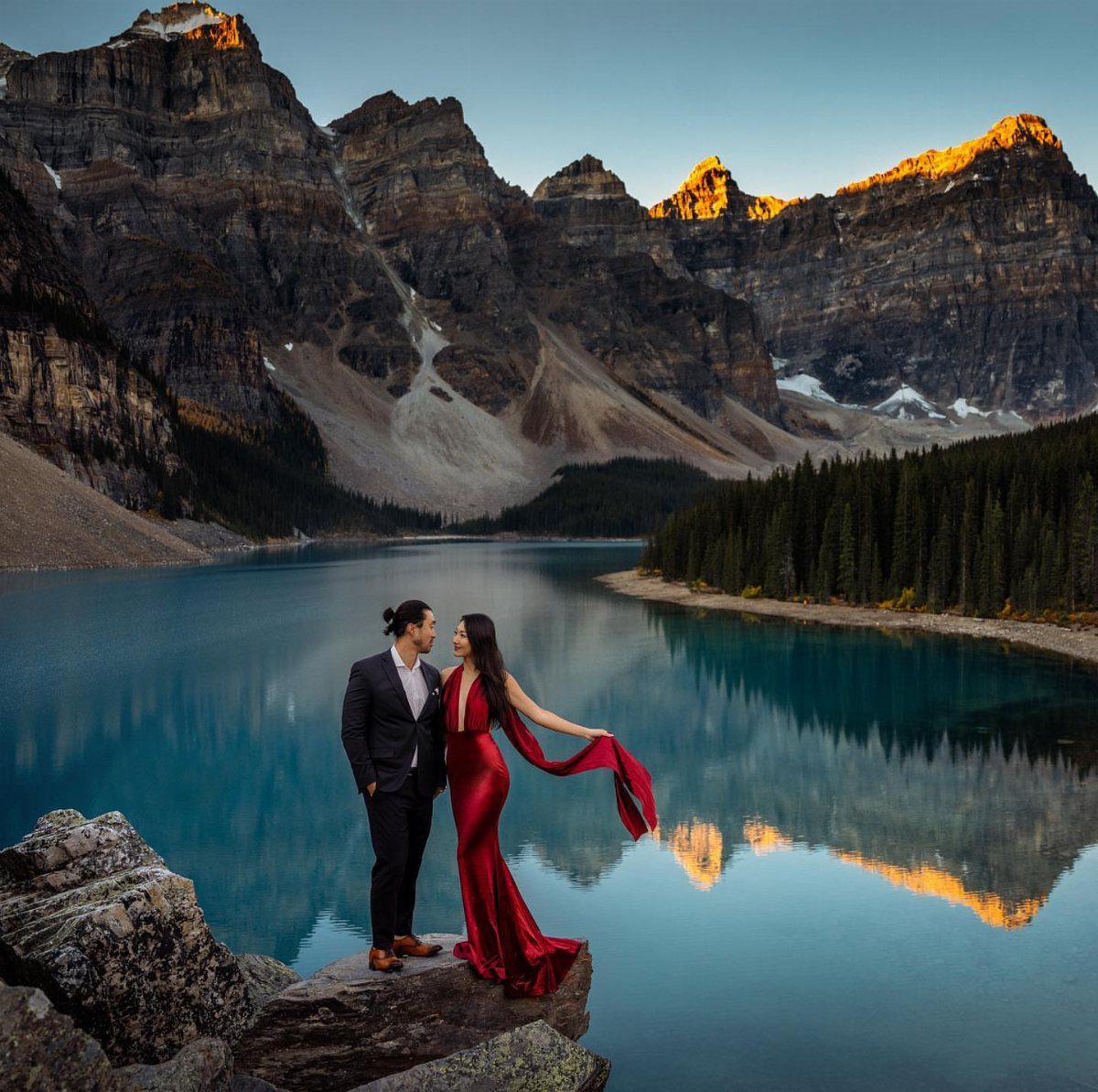 Couple in formal wear posing on a rocky cliff overlooking a mountain lake.