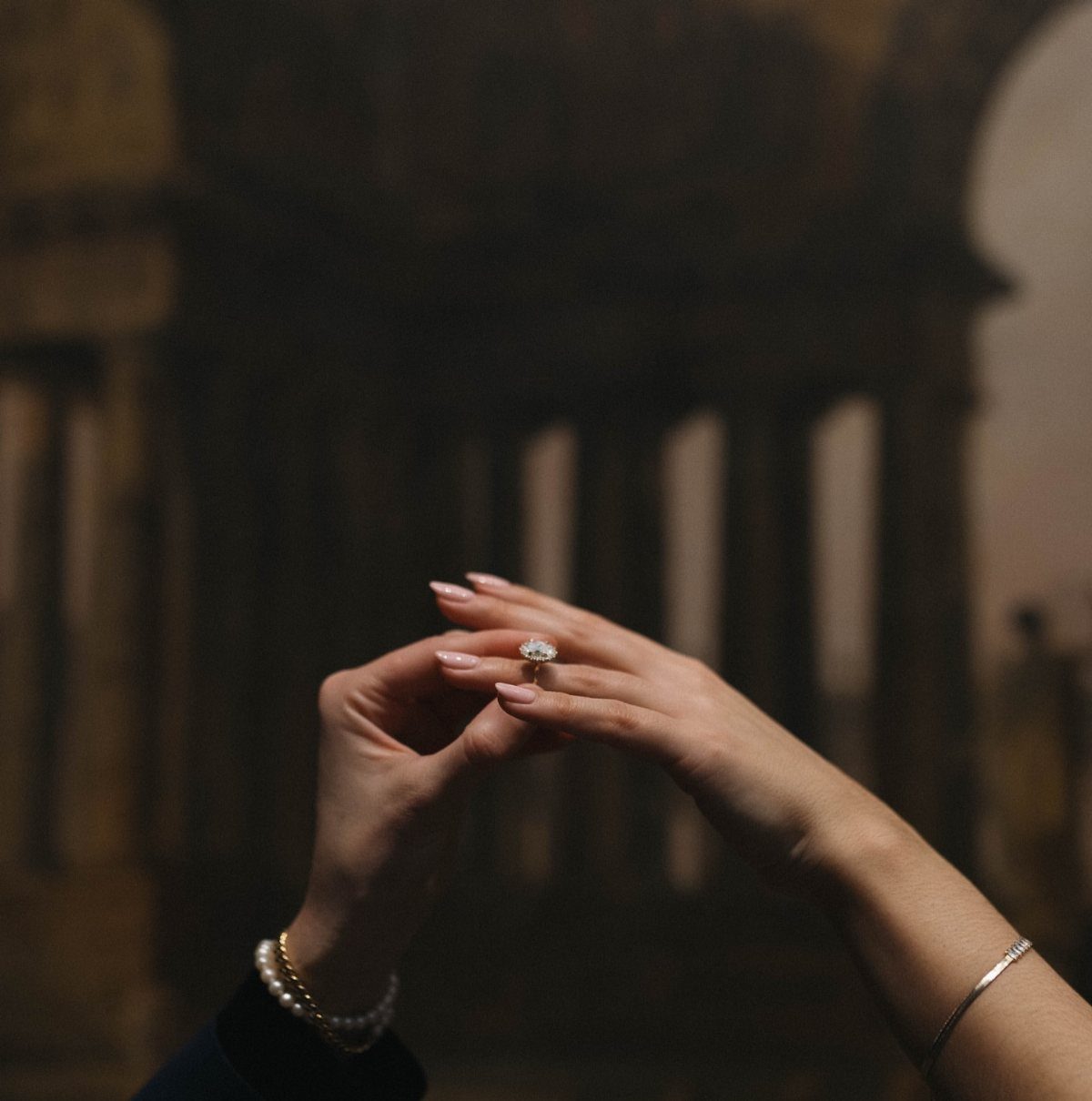 Hands holding a diamond engagement ring against a blurred architectural background.