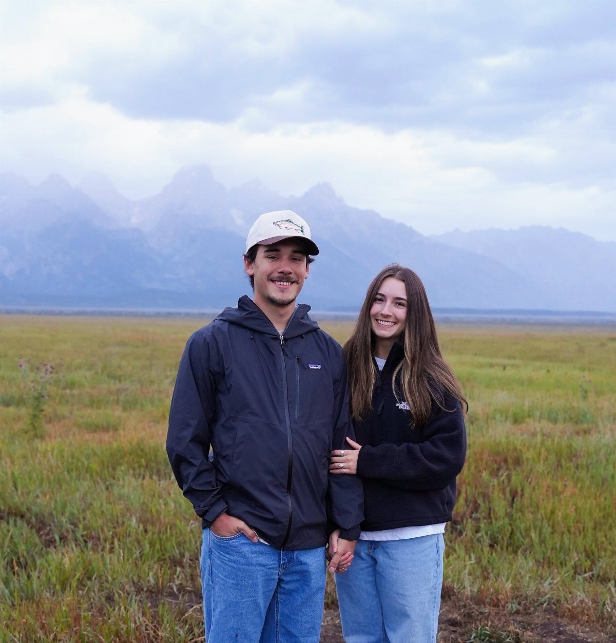 Young couple standing side-by-side in a grassy field with mountains in the background.