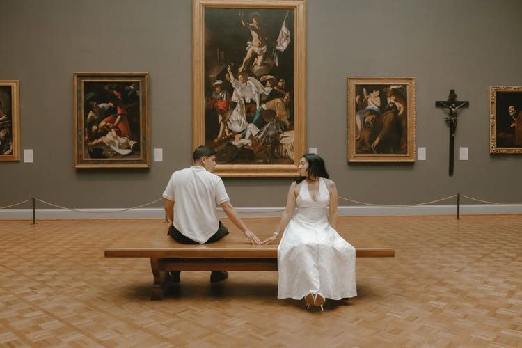 Couple in white attire sitting on a bench in an art gallery holding hands